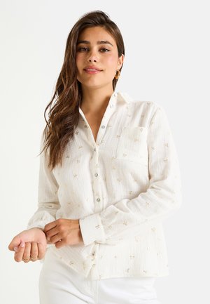 Chemise blanche à boutons avec de délicates broderies dorées, tissu texturé, col et une seule poche sur la poitrine ; le modèle pose avec les manches légèrement retroussées.