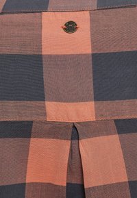 Rutigt skjorta med stora orange och marinblå rutor, med en plissédetalj på baksidan och en märkesmetallbadge nära kragen.