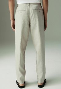 Pantaloni in lino grigio chiaro con vestibilità rilassata, dotati di tasche posteriori con zip e un tessuto testurizzato. Indossati con sandali neri.