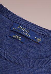 Pull en coton Pima bleu marine avec col côtelé. Les étiquettes indiquent "Polo Ralph Lauren", "Slim Fit" et les instructions d'entretien en texte jaune contrastant.