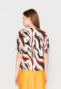 T-shirt de manga curta com um padrão de ondas coloridas em tons de castanho, rosa e laranja. Apresenta um corte solto e mangas ligeiramente bufantes.