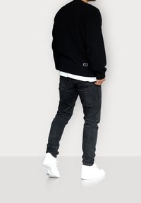 Pull noir en tricot avec ourlet côtelé, porté par-dessus une chemise blanche, associé à un jean noir et des baskets blanches. Vue de dos.