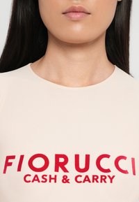 Svijetloružičasti prilagođeni top s odvažnim crvenim tekstom: "FIORUCCI CASH & CARRY." Glatka tekstura, okrugli izrez i kratki rukavi.
