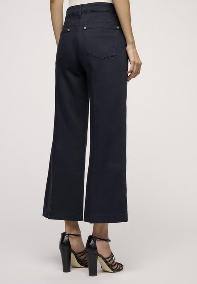 Jeans a gamba larga di un blu navy scuro con vita alta, dotati di due tasche posteriori e dettagli in metallo dorato. Il materiale ha una finitura liscia e leggermente texture.
