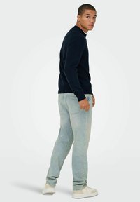 Maglione a maglia blu navy con polsini a coste, abbinato a jeans dritti di un lavaggio chiaro e sneakers bianche. Il modello sta in piedi di lato.
