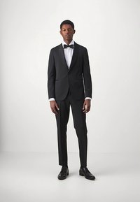 Mees seisab seljas must smoking, valge ülikonna särgiga, musta kikilipsu ja mustade kingadega, taustal on lihtne valge taust.