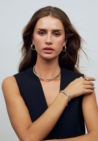 Sølv choker halskæde med et rørformet design, hoop øreringe og et struktureret armbånd båret på en bar arm; model i et sort outfit.