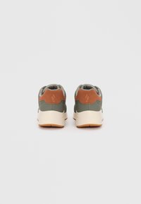 Sneakers verzi și bej cu o textură netedă, având un accent de tocul din piele întoarsă maro și o talpă din cauciuc alb. Vederea din spate arată detalii de umplutură.