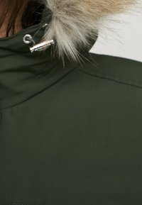 Dunkelgrüner Parka mit einer Kapuze aus Kunstfell, ausgestattet mit silbernen Beschlägen und einer glatten Oberfläche. Detail an der oberen Schulter sichtbar.