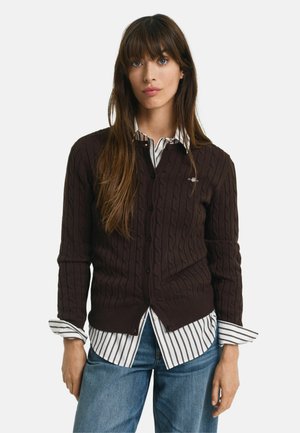 Brown cable-knit cardigan with a V-neck, button closure, and ribbed cuffs, layered over a white and gray striped shirt.