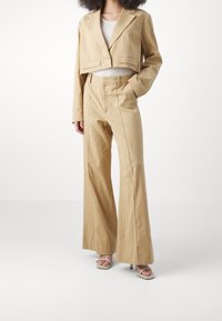 Beżowa krótka marynarka z dwoma przednimi kieszeniami, zestawiona z beżowymi szerokimi spodniami z wysokim stanem. Oba elementy wykonane są z tkaniny o fakturze.