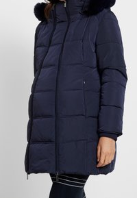 Navy puffer jacket with a high collar and fur trim, featuring a quilted design, front zippers, and ribbed cuffs. Long, curved silhouette.