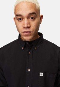 Sort button-up skjorte lavet af bomuld, med krave, træknapper og en brystlomme med et logo på venstre bryst.