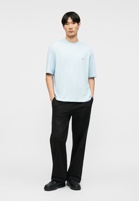 Νεαρός άνδρας στέκεται με τα χέρια στις τσέπες, φορώντας ένα ανοιχτό γαλάζιο φαρδύ μπλουζάκι, μαύρο φαρδύ παντελόνι και μαύρα παπούτσια σε λευκό φόντο.