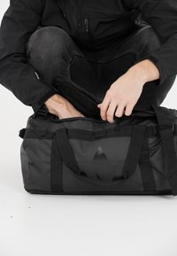 Musta urheilukassi tukevilla kahvoilla, kiiltävästä materiaalista, suorakulmainen muoto ja teksturoitu sisus. Sisältää hienovaraisen graafisen suunnitteluelementin.