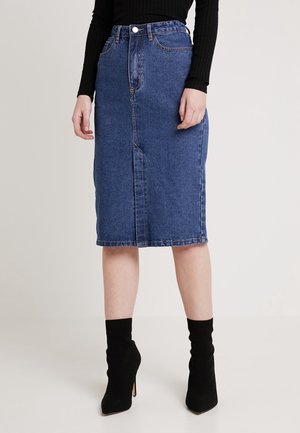 Mujer que lleva una falda vaquera azul hasta la rodilla, top negro de manga larga y botines negros con tacón, de pie frente a un fondo liso.