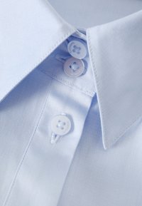 Camicia azzurro chiaro con colletto a punta, due bottoni bianchi con motivo floreale e una trama tessile liscia.
