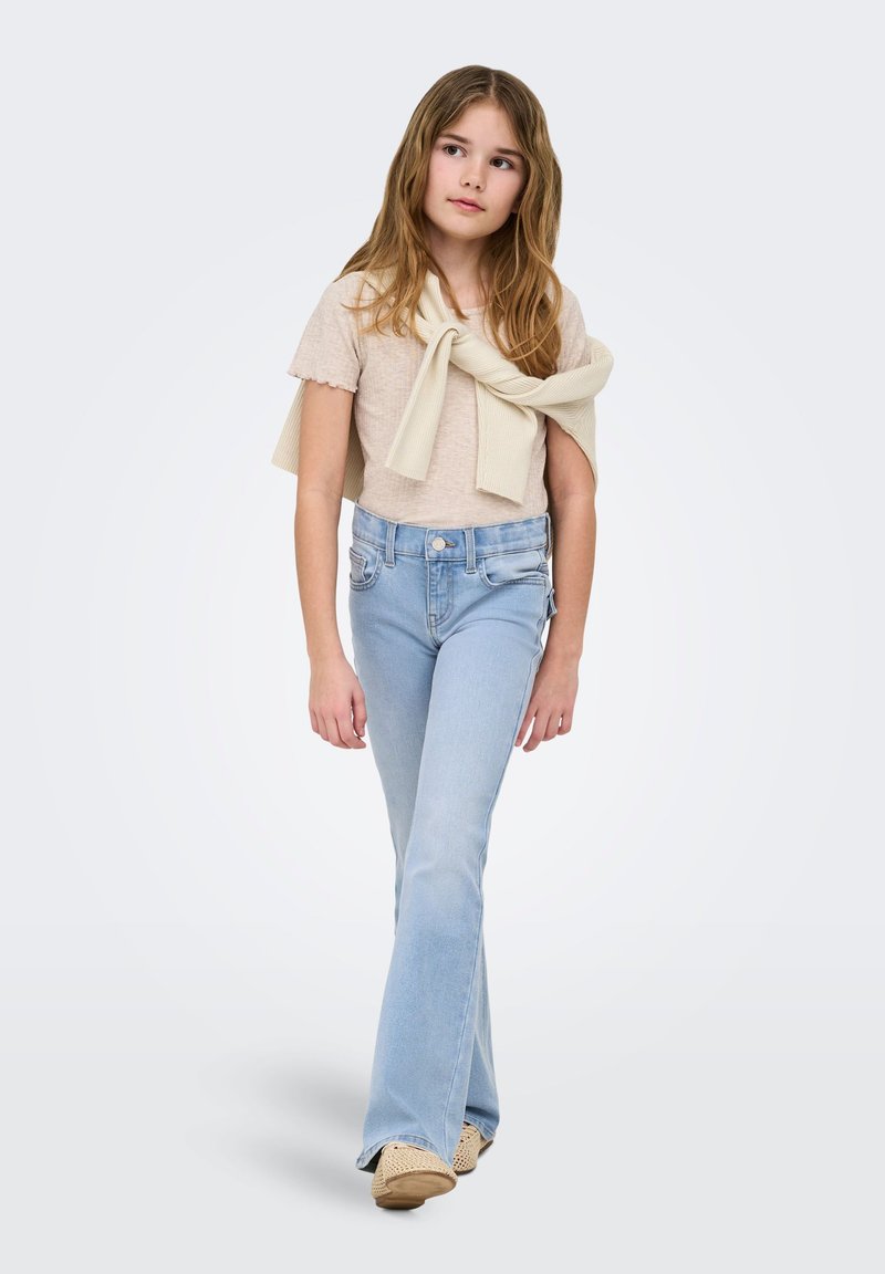 Ragazza giovane che cammina, indossa una maglietta beige chiaro, un maglione crema legato sulle spalle, jeans a zampa chiaro e scarpe beige intrecciate.