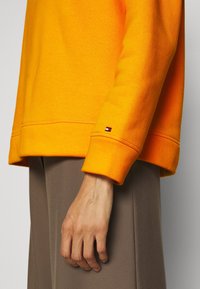 Sudadera naranja con un corte holgado, puños y dobladillo acanalados. Etiqueta de logo pequeña en la manga. Combinada con pantalones marrones. Textura suave.