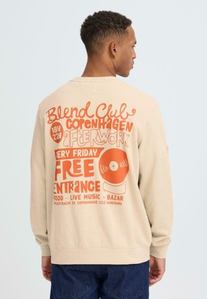Beige Sweatshirt mit lockerem Sitz, auf der Rückseite mit auffälligem orangem Text zur Bewerbung einer Veranstaltung, mit gerippten Bündchen und Saum.