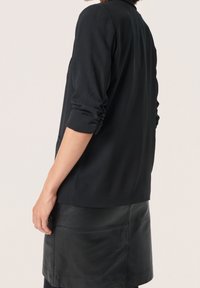 Giacca nera con una texture morbida, con maniche arrotolate e una vestibilità rilassata. Indossata sopra una gonna in pelle nera aderente.