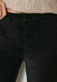 Primo piano di una persona che indossa pantaloni di velluto a coste neri e un maglione corto lavorato a maglia beige, con una mano poggiata vicino alla vita.