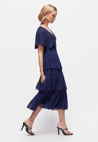 Vestido azul marino de capas con un diseño en forma de concha, cuerpo adornado y una superposición transparente. Combinado con sandalias negras de tacón y pendientes de botón.