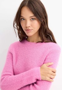 Joven con cabello largo castaño que lleva un suéter de lana de color rosa brillante, sosteniéndose el brazo, con un fondo neutro.