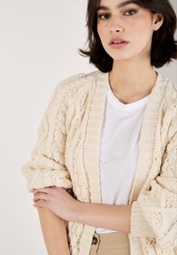 Fleckige Creme-Strickjacke mit Kabelmustern, offenen Vorderteilen und weiten Ärmeln über einem weißen T-Shirt. Minimalistisches Design mit strukturiertem Stoff.