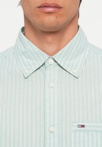 Hellgrün kurzärmliges Hemd mit weißen vertikalen Streifen, Button-Down-Kragen und einer Brusttasche mit einem Logo-Patch.