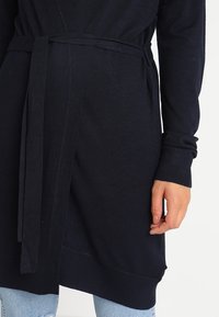 Granatowy sweter z dzianiny z paskiem do wiązania, prążkowanymi mankietami i fakturowanym materiałem. Sweter ma luźny krój i sięga do połowy uda.