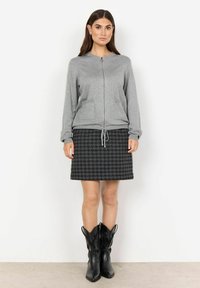 Grå dragkedja sweatshirt med fickor, tillsammans med en svart kjol i houndstooth-mönster och svarta cowboyboots. Slätt tyg och strukturerad design.