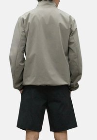 Chaqueta impermeable de color gris claro con un alto cuello, puños elásticos y un corte holgado, combinada con pantalones cortos negros. Textura suave y mate.