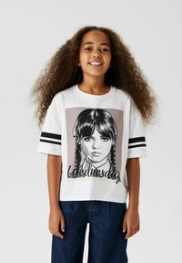 Vit t-shirt med kort ärm som har en illustration av en flicka med flätor. Texten nedan lyder "Onsdag" och "Var den konstiga du vill vara."