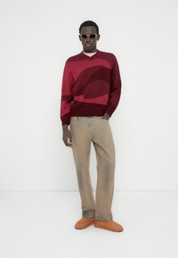 Μπορντό πλεκτό πουλόβερ με κυματιστά σχέδια, συνδυασμένο με μπεζ τζιν φαρδιάς γραμμής και καφέ παπούτσια χωρίς κορδόνια. Αξεσουάρ περιλαμβάνουν στρογγυλά γυαλιά ηλίου.