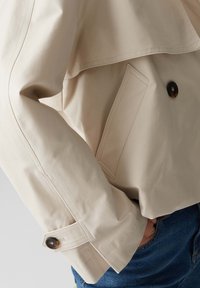 Giacca beige realizzata in tessuto liscio, con un fronte doppiopetto e bottoni scuri, e colletto a bavero. Dettaglio con tasche laterali visibili.