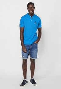 Modrá polo košile s knoflíkovým límečkem a krátkým rukávem; v kombinaci s džíny v odstínu denim s opotřebovaným vzhledem. Nosí se s tmavými teniskami.