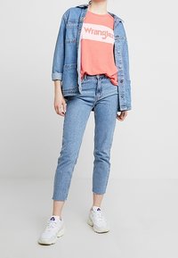 Denimjakke over en koralfarvet t-shirt med "Wrangler" logo, parret med lys blå skinny jeans og hvide sneakers. Enkel, afslappet outfit.