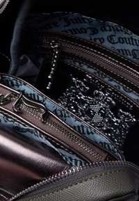 Interno in bronzo metallico con tasche con zip, dotato di una fodera in tessuto blu, accentuata da testo nero. L'esterno è texturizzato in verde oliva.