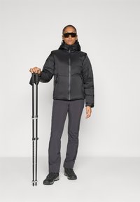 Femme portant une doudoune noire, un pantalon sombre, des bottes de randonnée et des lunettes de soleil, tenant deux bâtons de trekking noirs devant un fond uni.