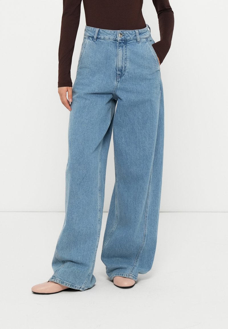 Jeans de mezclilla de tiro alto y pierna ancha en azul claro, con cierre de botón en la parte delantera y bolsillos laterales, con una textura suave y ligeramente deslavada.