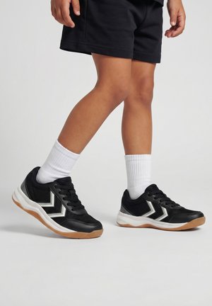 Dziecko noszące czarne buty sportowe z białymi logo w kształcie chevron, białe skarpetki i czarne spodenki, stojące na jasno szarej podłodze.