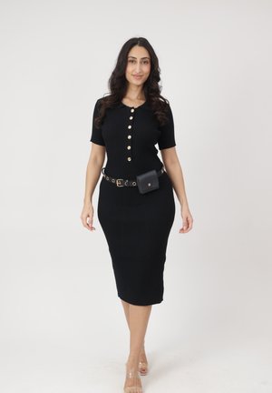 Frau in einem enganliegenden schwarzen Midikleid mit goldenen Knöpfen, einer schwarzen Bauchtasche und klaren Riemchenabsätzen, die vor einem schlichten hellen Hintergrund steht.