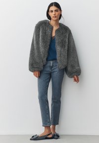 Woman wearing a gray faux fur jacket, teal lace-trimmed top, blue jeans, and black flat shoes with rhinestone bows, standing against a white wall.