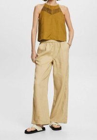 Top halter de mostaza con pliegues horizontales, combinado con pantalones de pierna ancha beige. La modelo lleva sandalias de color crema con suelas negras.