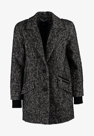Manteau oversize en pied-de-poule noir et blanc, chemise noire, jean bleu, sac fourre-tout noir et chaussures noires à bouts pointus.