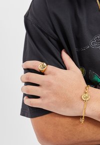 Gold ring with intricate design on the hand, paired with a gold chain bracelet featuring a circular charm, worn over a black shirt.