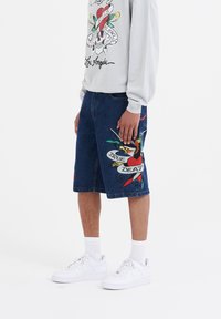 Los pantalones cortos de mezclilla con un ajuste relajado cuentan con un bordado inspirado en tatuajes de un pájaro y un corazón, que muestran detalles vibrantes en rojo, verde y azul.
