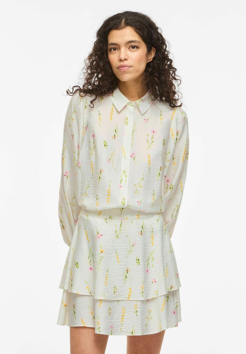 Femme aux cheveux bouclés portant un chemisier blanc à col et une jupe assortie à couches avec de petites imprimés floraux jaunes et roses.