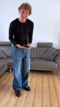 Svart långärmad skjorta, blå vida jeans, bruna skor. Person håller i en svart sak, står på trägolv bredvid en grå soffa.